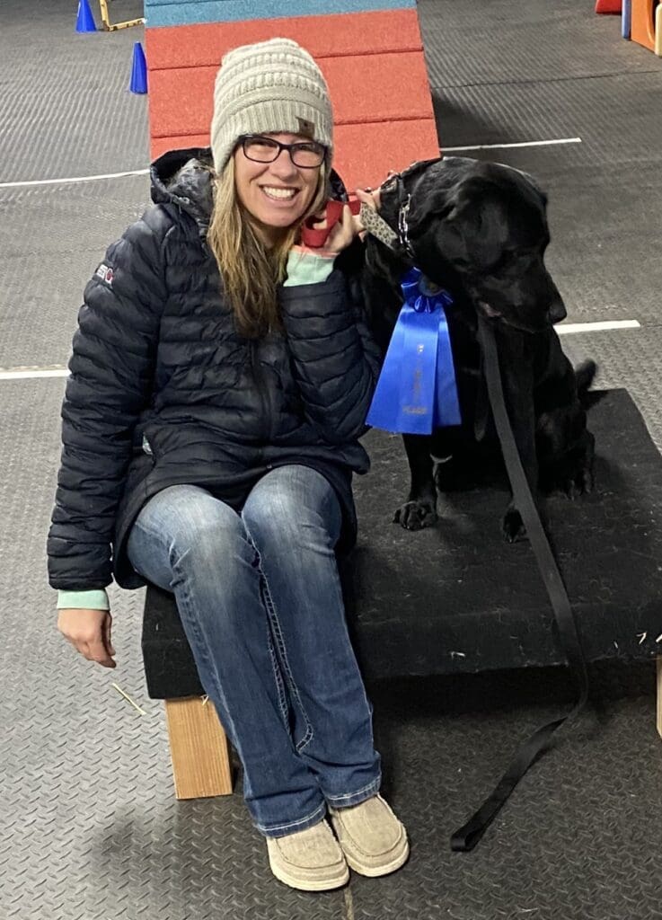 Melissa sitting beside Olive during an agility training event, Both bundled up and smiling.