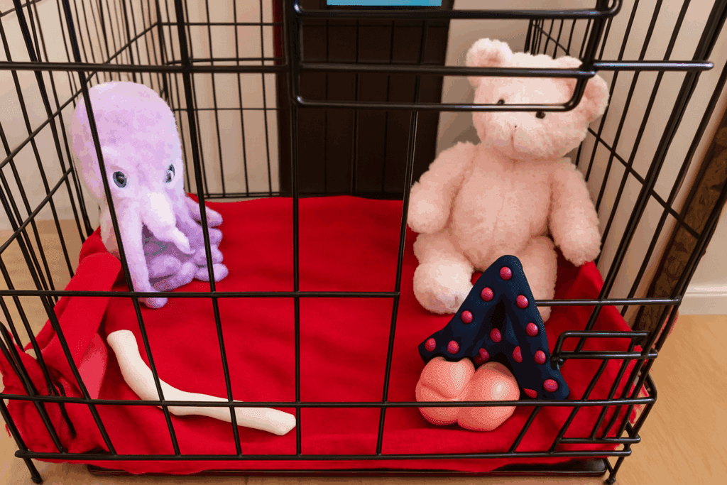 Open dog crate with a red blanket, plush octopus toy, pink teddy bear, and a puppy teething bone arranged inside.