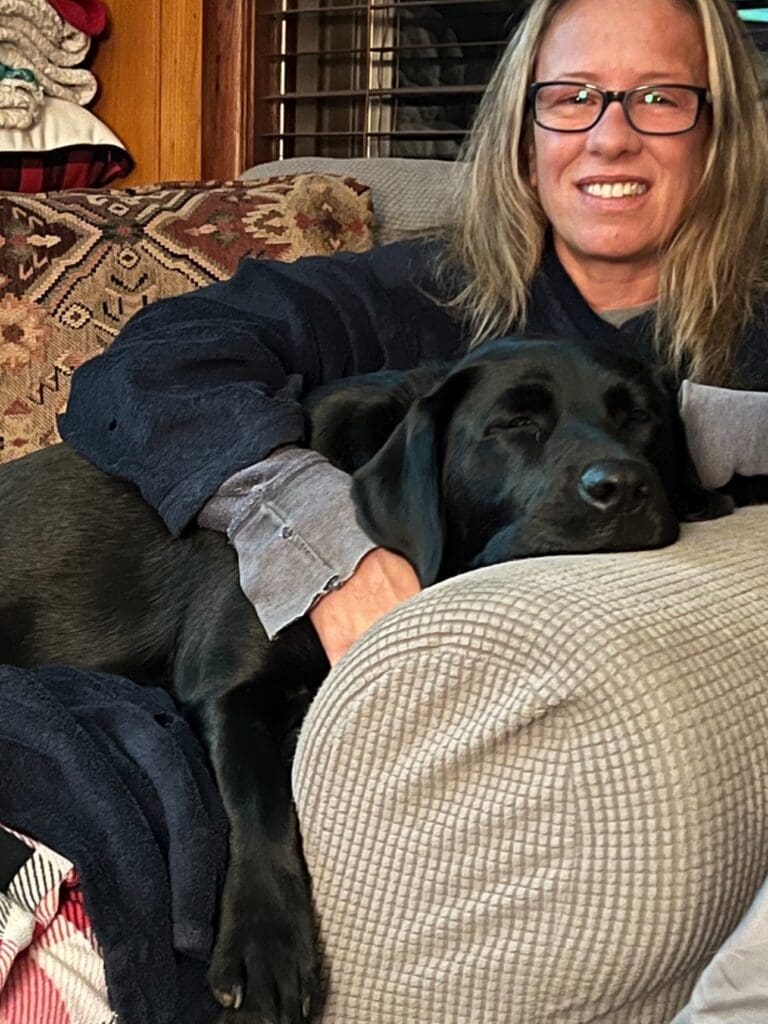 Olive the black Labradore cuddled up in the chair with Melissa, both look cozy indoors.