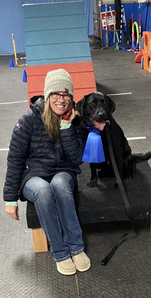 Melissa sitting beside Olive at an indoor agility course, both smiling after training.